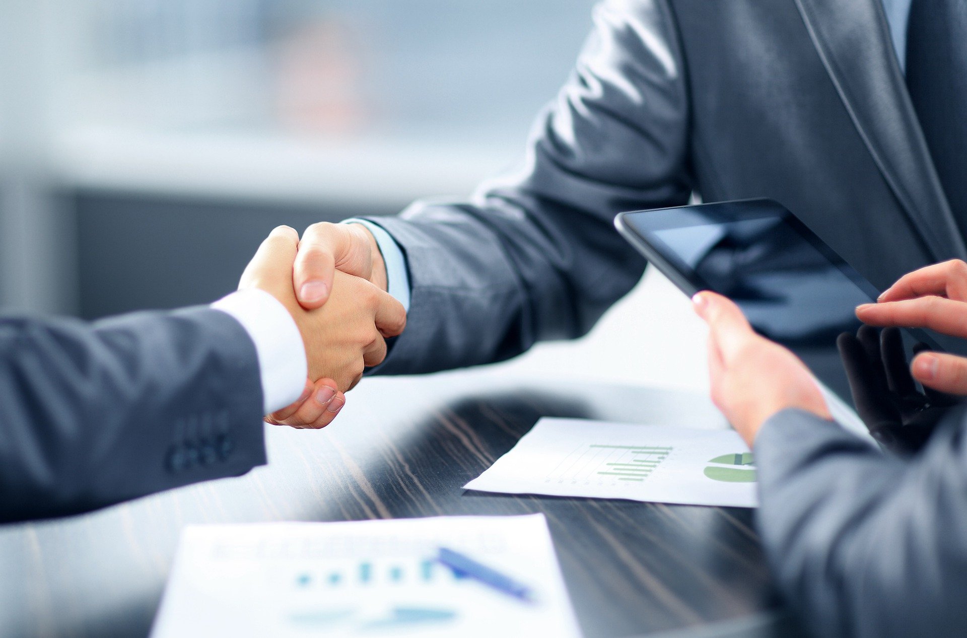Two men in suits shaking hands after completing a deal. You only see their right arms and hands. There's a third man's hand on the right side of the image holding a phone.