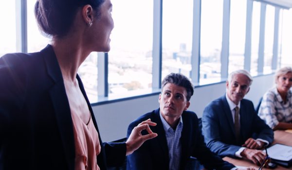 A room of business people at a table in a meeting. They're all seated on the same side of the table, looking at a woman who is standing at the head of the table and talking. Everyone else is seated. There are high windows in the background letting in a lot of natural light.