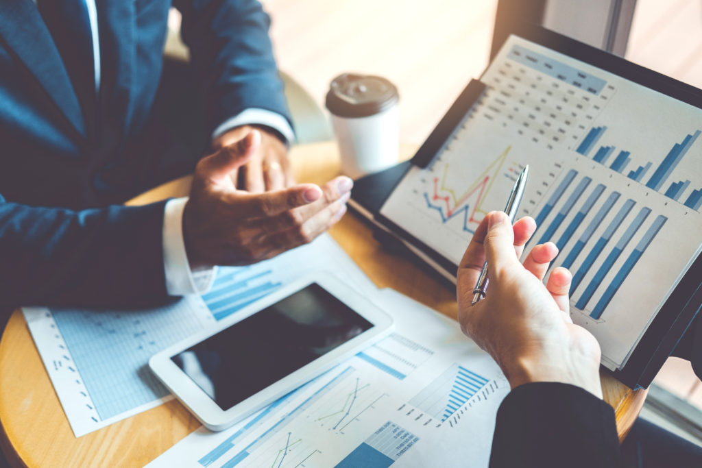 Two people discussing financial reports. There's a phone, a tablet and papers on the table. The papers and tablet show charts and data. One person is a man dressed in a suit. You only see the left hand of the other person. They're holding a pen. ot