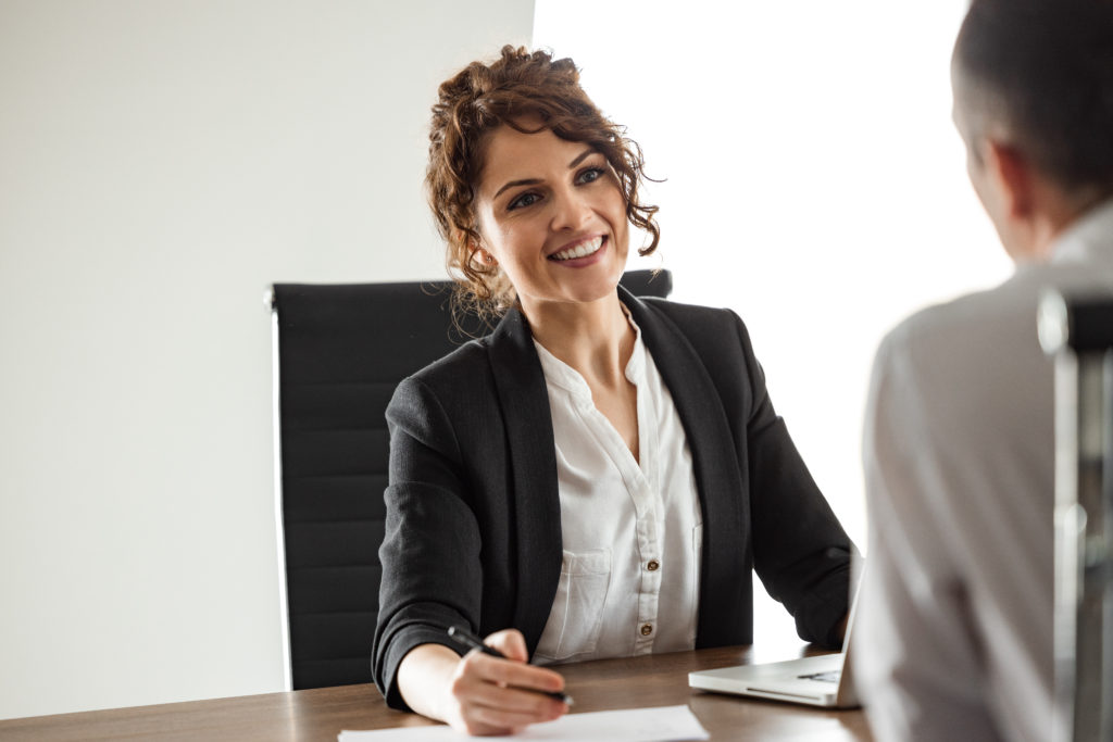 A businesswoman sitting in a chair at a desk talking to someone across the table from her