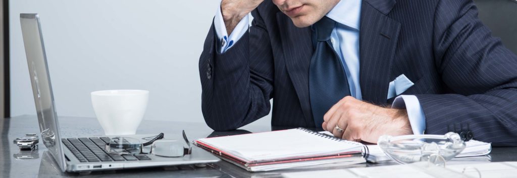 A man wearing a suit sitting at a desk with an open gray laptop, a white coffee mug and papers and folders. He's looking down and the desk and looks very stressed.