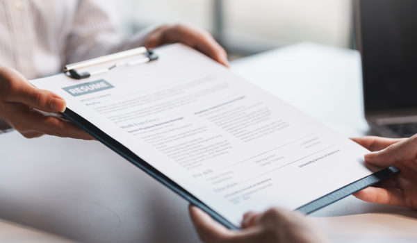 A resume attached to a clipboard is being handed over a desk from one person to another