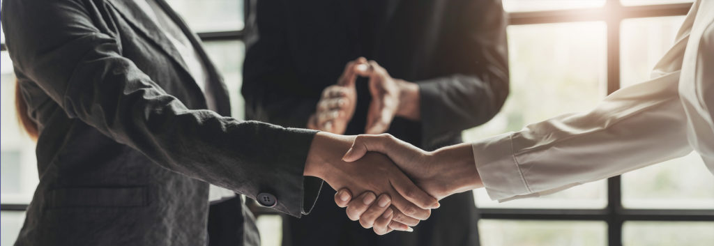 Two people are shaking hands while someone else excitedly observes in the background. You can only see from above the waist to below the neck for all three people.