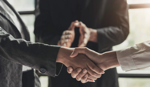 Two people are shaking hands while someone else excitedly observes in the background. You can only see from above the waist to below the neck for all three people.