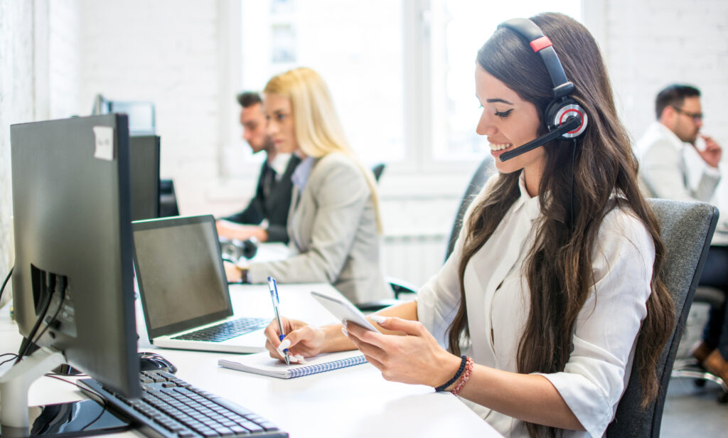 Friendly customer support female operator with headphones writing notes to notebook taken from smartphone at office