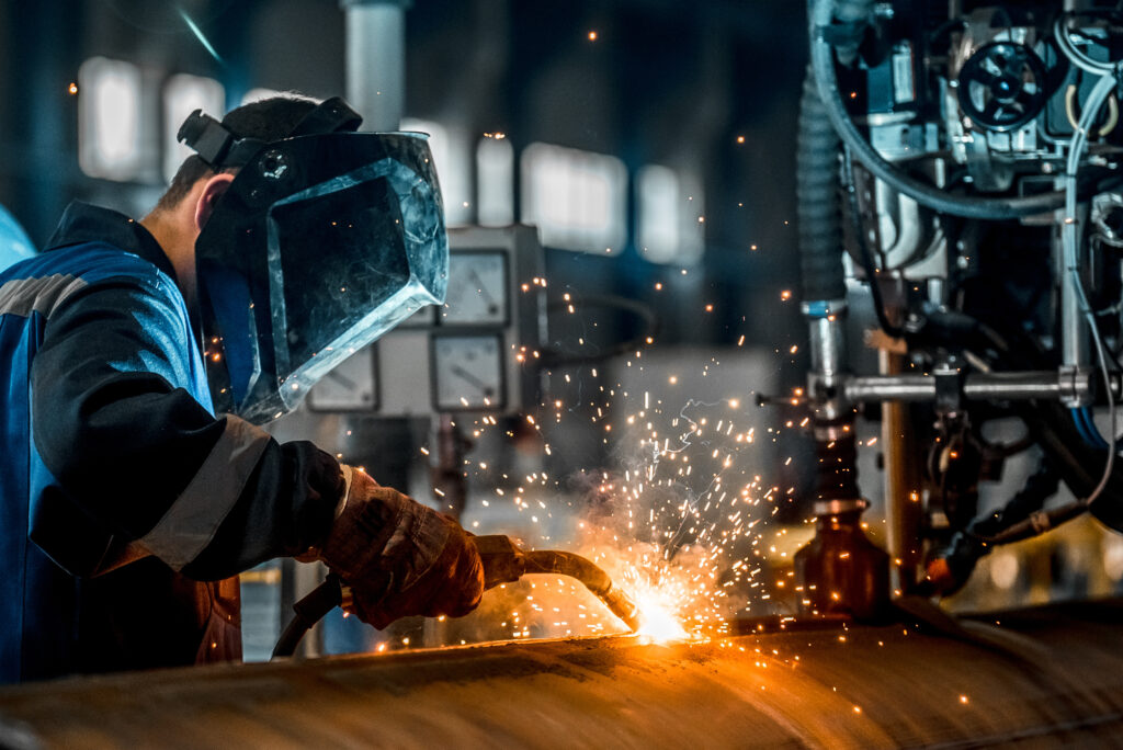 Man welds at the factory with a protective helmet on. It's pretty dark. The only light comes from the sparks he's creating while welding.