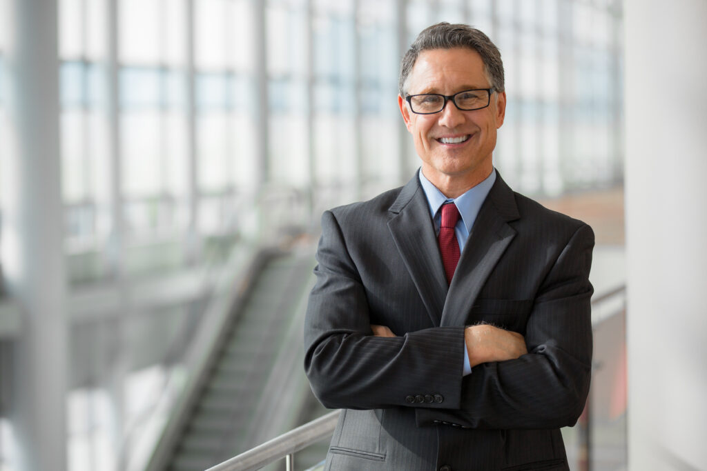 A man in a suit is smiling and standing with his arms crossed. In the background is a big open lobby with very high glass windows and the top half of an escalator.