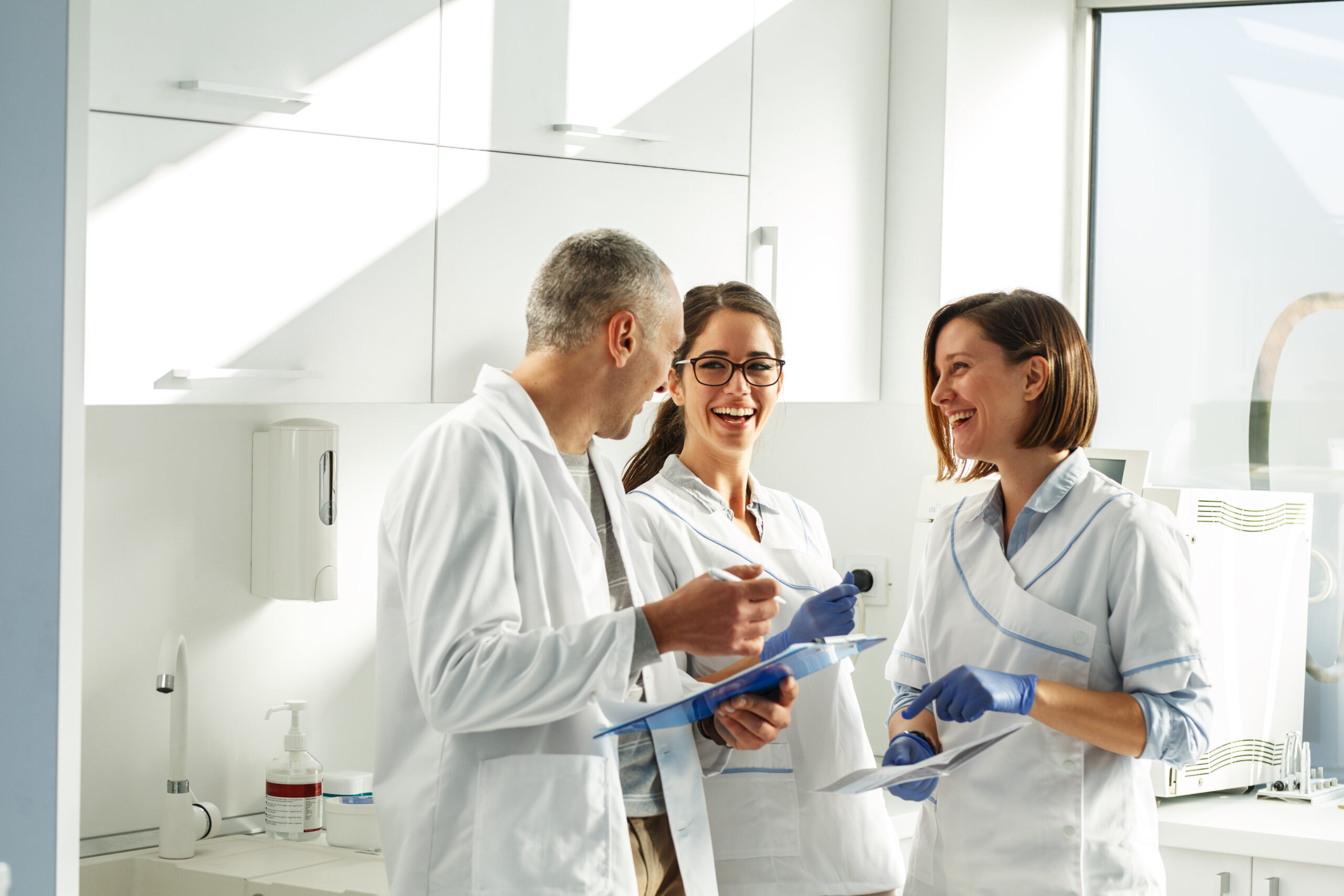 Three dentists – one male and two females, all white – stand in a well-list dentist office smiling. The room is almost completely white and has a large window on the right.