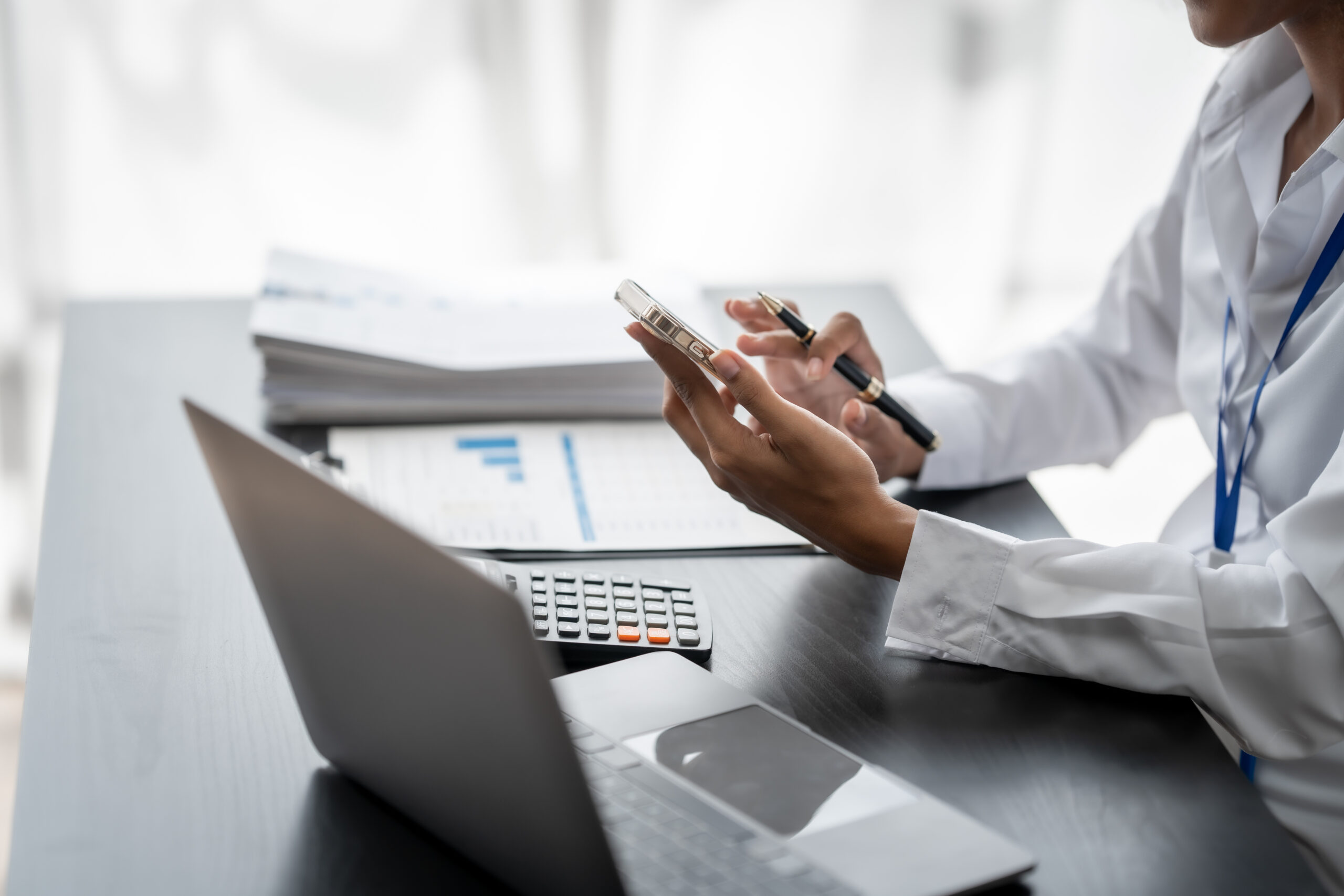 A black business woman's hands shown holding a phone with a calculator, gray laptop and two stacks/piles of papers on the desk in front of her