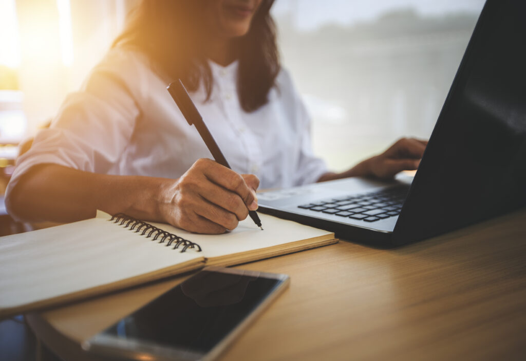 A woman at a desk with a computer. You can't see the top half of her face. She's wearing a white shirt while writing something in a notebook to the right of the computer. Her cell phone is also on her desk. It appears to be sunrise or sunset in the background.