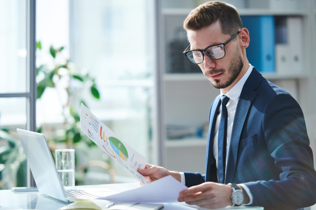 Serious young economist in eyeglasses and formalwear looking through financial papers by workplace