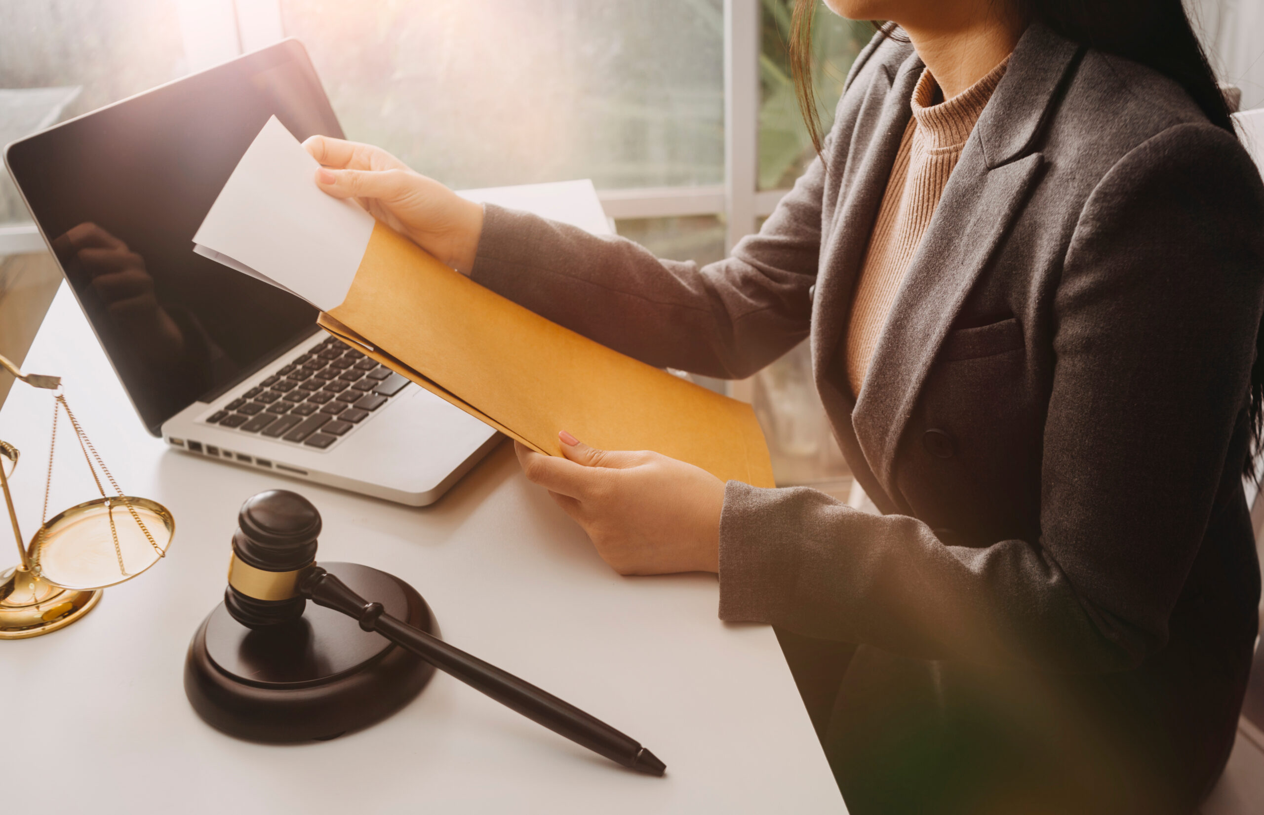 A lawyer taking papers out of a manila envelope. There's a gavel block on the desk to the left of a laptop computer. It appears to be late afternoon or early morning based on the natural light coming from the person's right. You don't see their head.