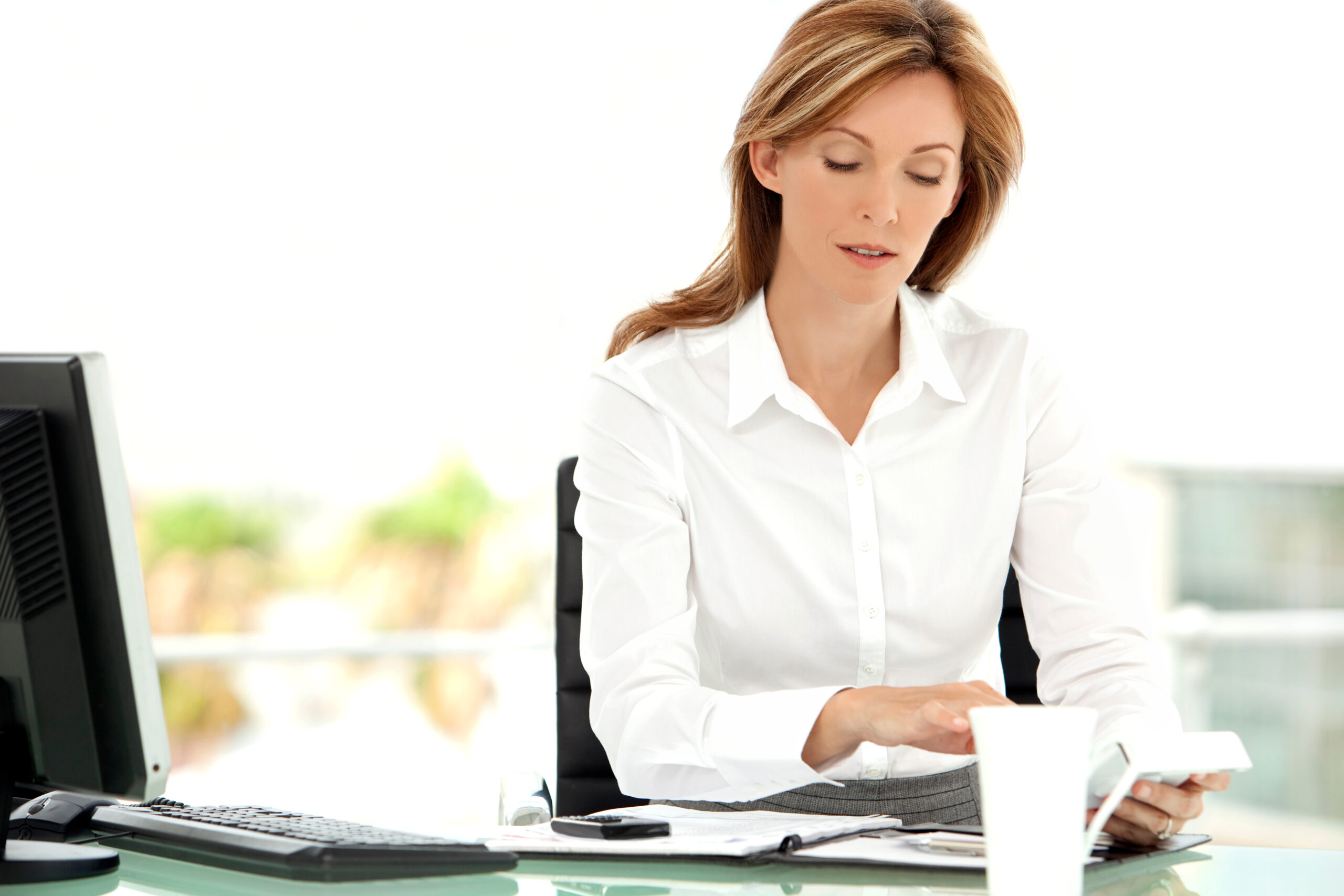 A dirty-blonde white woman in a white button-up is working at a desk with a serious look on her face. There's a mug on the desk, too. The background is well-lit with natural light.