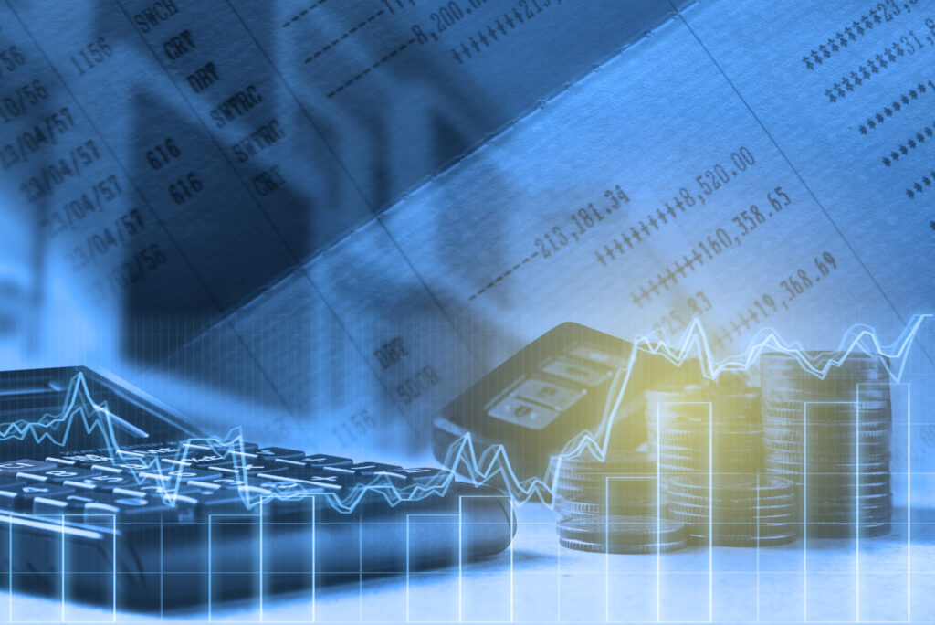 A calculator and a stack of coins in a bluish tinted photo. In the background there are accounting papers with numbers on them. In the foreground there's a bar chart with a line graph.