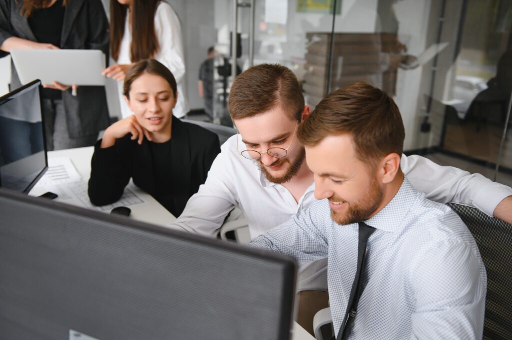 Three salespeople are seated in front a couple monitors, but they're looking at something on the desk. There are two males and a female, all white. The middle person is a male wearing glasses. Both males have beards. One has a tie and the other just a button-up that's open at the top. The woman has her right elbow on the table and is wearing all black. There are two more women standing in the background looking at a laptop. They're all in an open office with glass walls and doors.