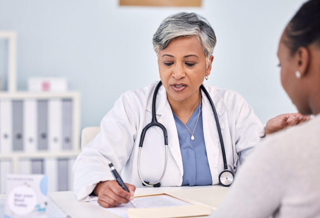 A black female doctor with peppered hair sits across the table from a black female patient who has an earring in her left ear. The doctors has a stethoscope hanging around her neck and is writing something with a black click pen in her right hand. The background is blurred, but there's a robin-egg blue wall and some other objects, possibly binders.