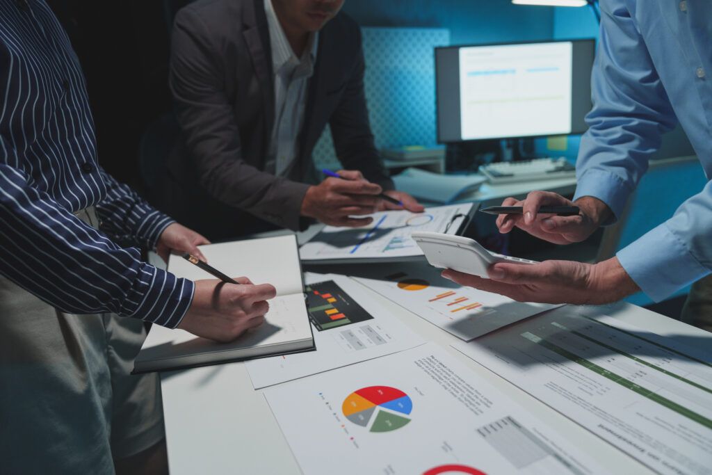 Three people are around a table full of papers that feature pie charts and bar graphs. Each of them has a pen in one hand. Each has something the other hand – a calculator, a notebook, a clipboard holding a paper with more charts. There's a computer screen flowing in the background. The room is lit by fluorescent lighting. All the people appear to be white, but you can only see half of one of their faces.