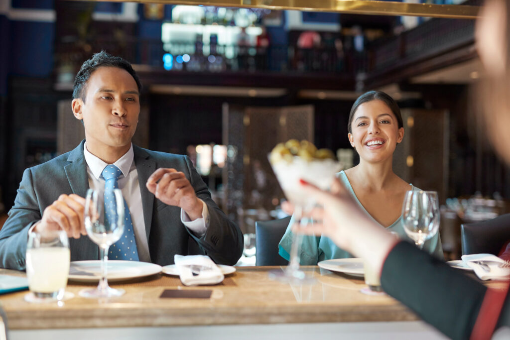 A man and a woman sit at a bar. Their plates and wine glasses are empty. The man's wearing a suit and tie while the woman is wearing an aqua dress. They're both tan. The man is probably of southeast Asian descent, and the woman Latina or middle-eastern. In the blurred foreground, a white female bartender's is passing a cocktail to the man with her right hand, which has bright fingernail polish. Her sleeve is black.