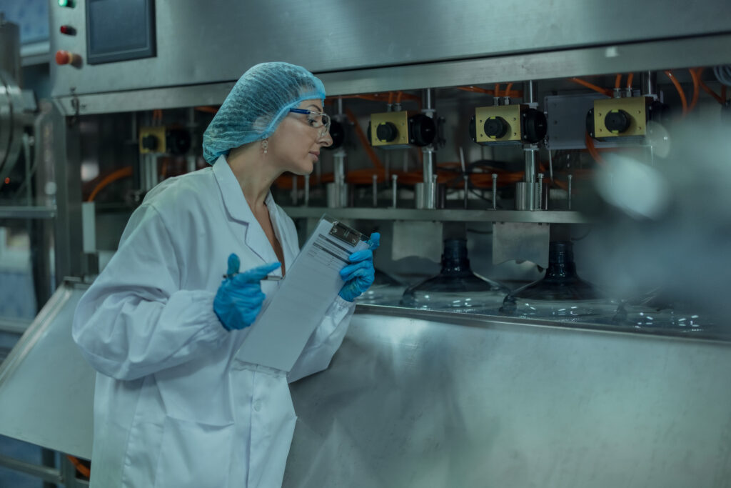 A female quality assurance specialist in a white coat, blue gloves and a blue hairnet is at a water production plant examining and recording information about conveyor operations to ensure proper timing and uninterrupted flow throughout the manufacturing process.
